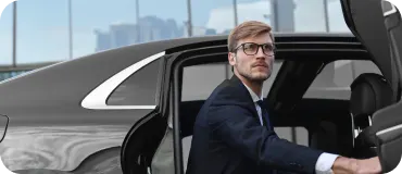 A man in a navy suit opening the door of a black sedan with a city skyline behind him.